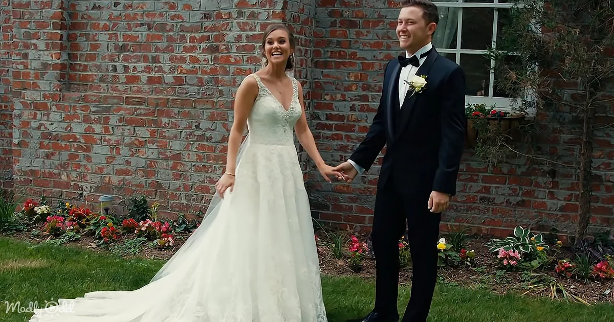 A bride in a white gown and a groom in a black tuxedo walk hand in hand in front of a brick wall with flowers.