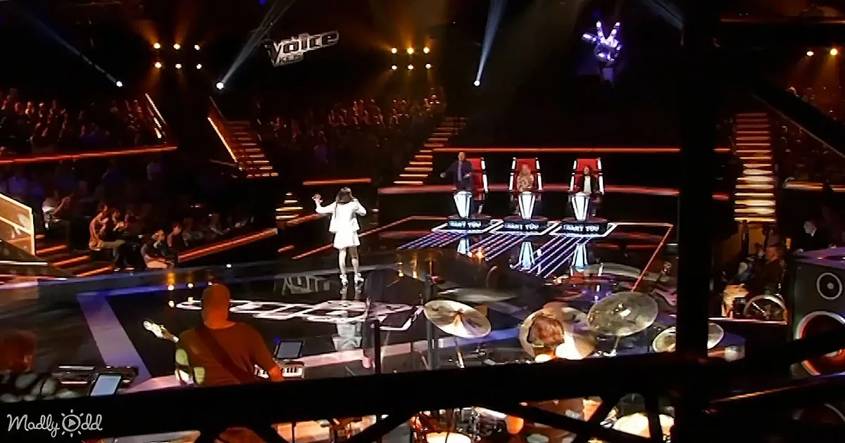 A performer in white stands on stage, surrounded by judges' chairs and a live band, with audience and stage lights in the...
