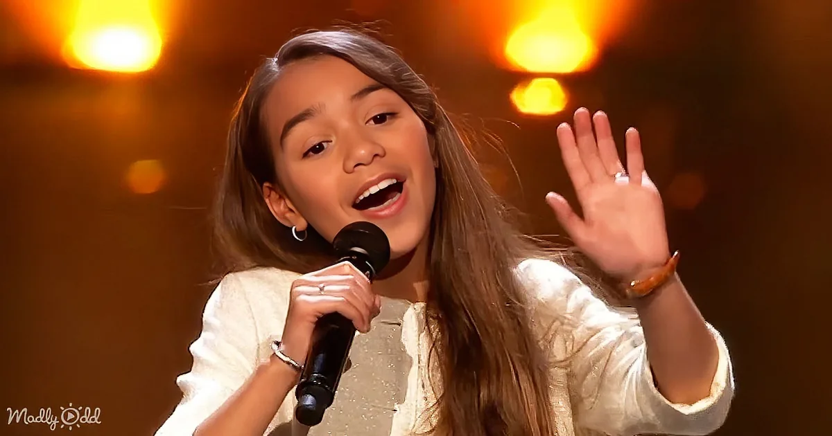 A young girl sings passionately into a microphone, raising her hand, with warm stage lighting behind her.