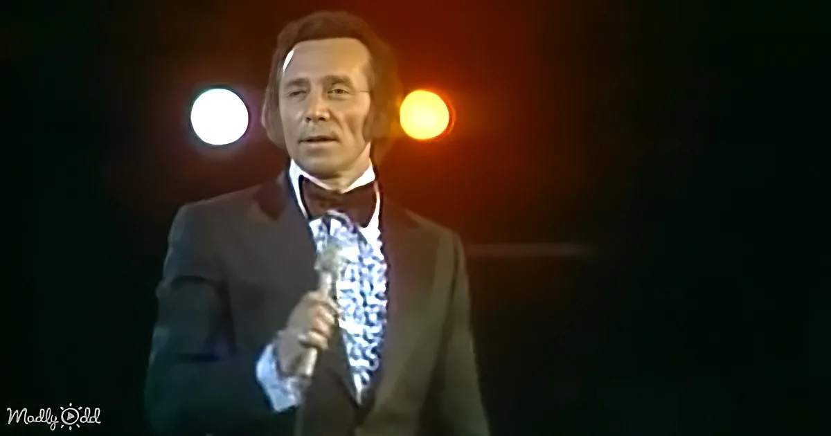 A man in a suit and bow tie holds a microphone on stage with bright lights behind him.