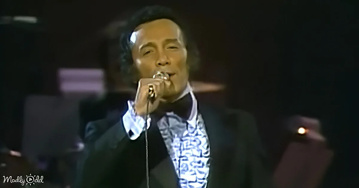 A man in a suit sings into a microphone on stage, wearing a patterned shirt and bow tie.