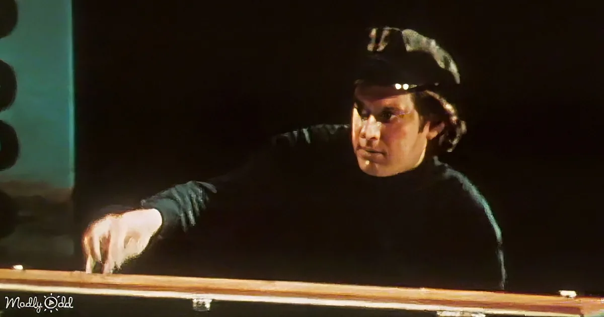 A man in a dark turtleneck and cap leans forward, appearing focused or concerned, in a dimly lit setting.