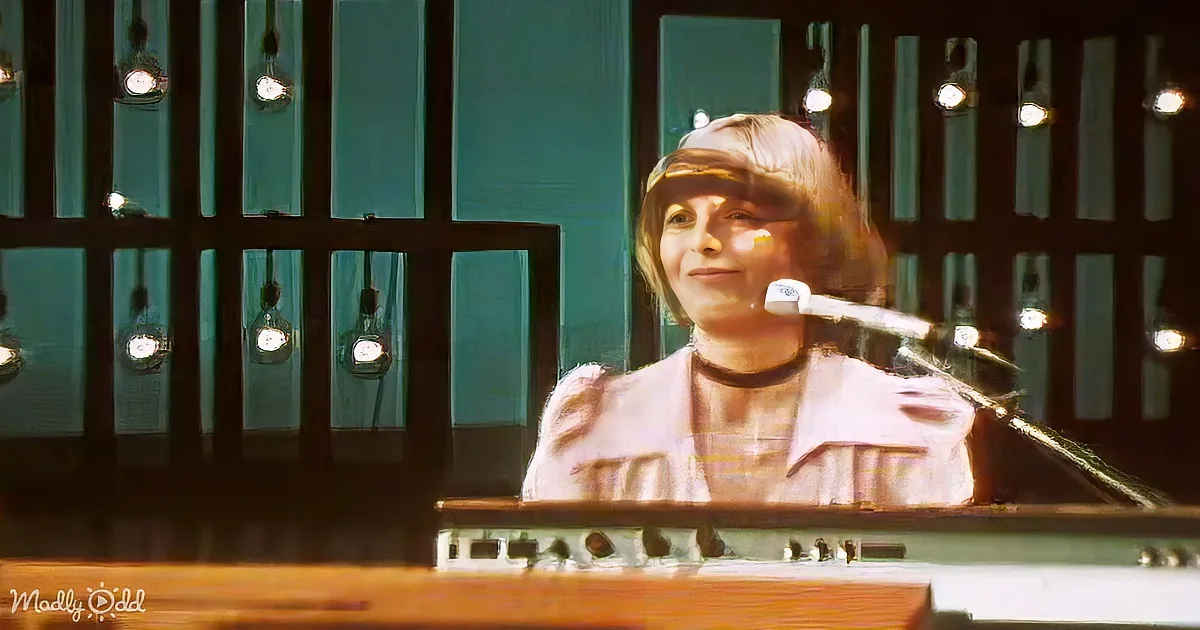 A person with short hair and a white jacket is sitting in front of a keyboard, surrounded by bright lights.