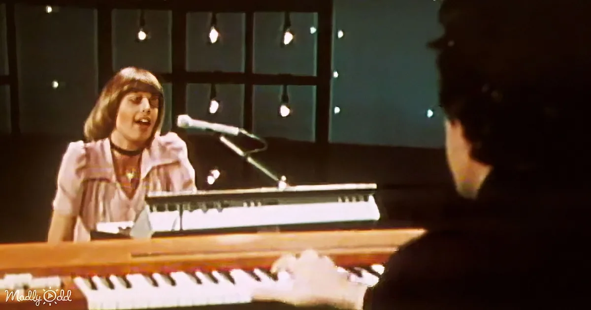A woman sings into a microphone while playing a keyboard, with another person playing a piano in the foreground.