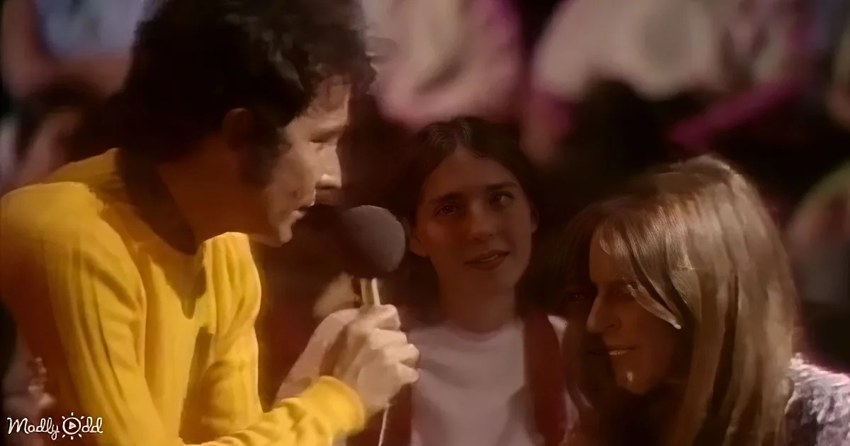 A man in a yellow shirt holds a microphone, singing or speaking to a smiling audience.
