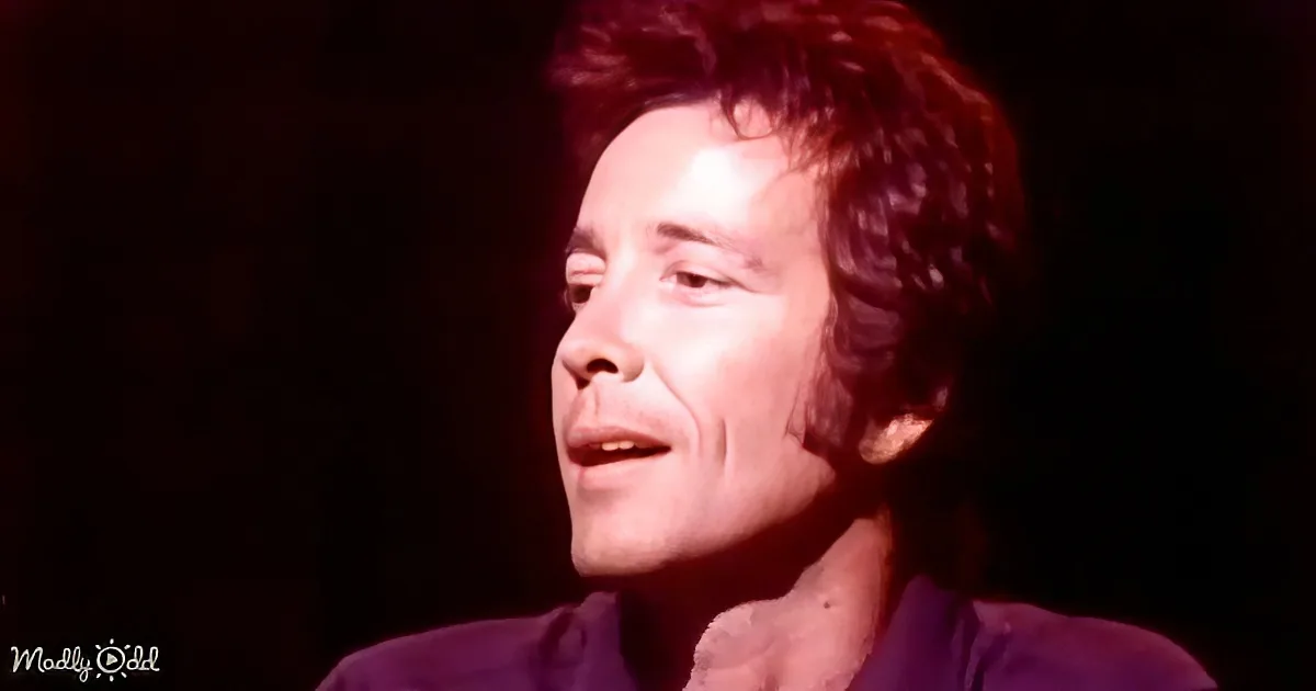 A person with curly hair and a purple outfit, illuminated by red lighting, appears to be speaking or singing.