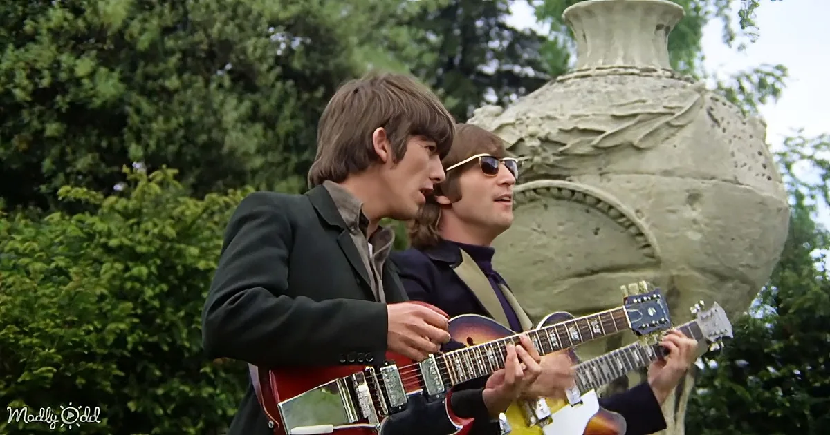 Two musicians play guitars outdoors, one singing, surrounded by greenery and a large stone sculpture.