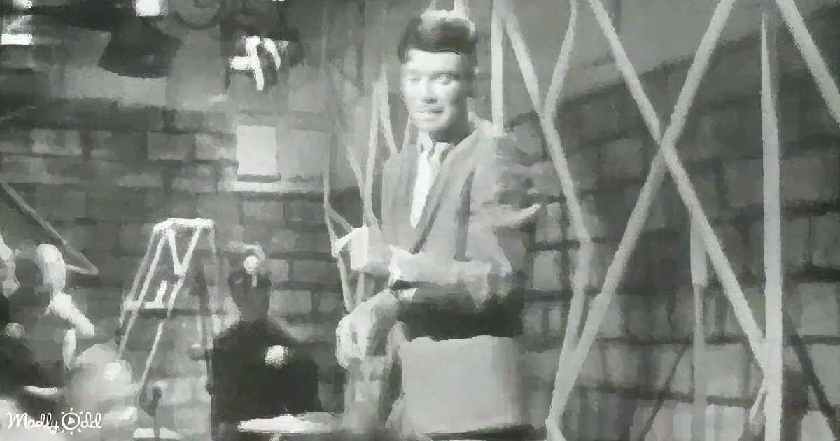 A man in a suit sits at a table, surrounded by scaffolding and a brick wall, with a microphone in front of him.