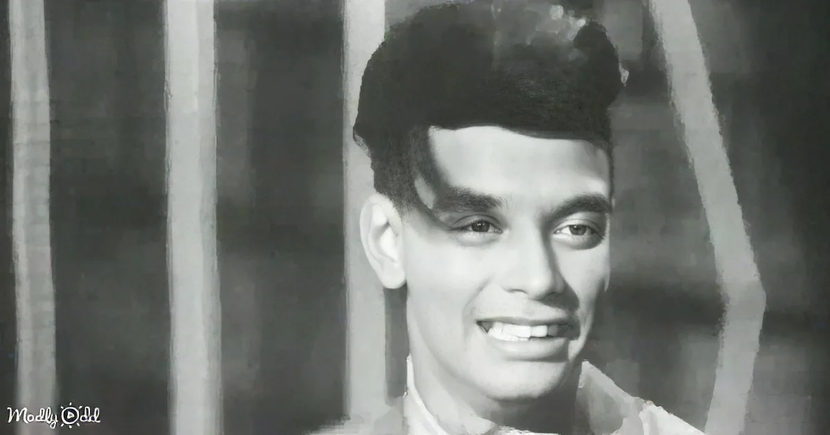 A smiling man with dark hair and glasses, wearing a light-colored shirt, stands in front of vertical bars.