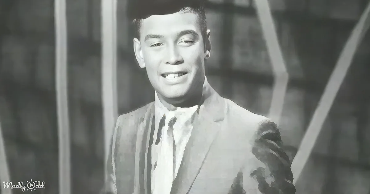 A smiling man in a suit and tie stands in front of a window, looking directly at the camera.