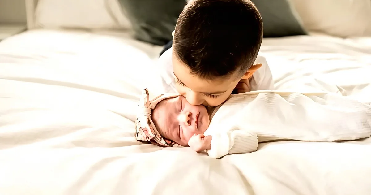 A young boy gently kisses a sleeping baby on a white bed.