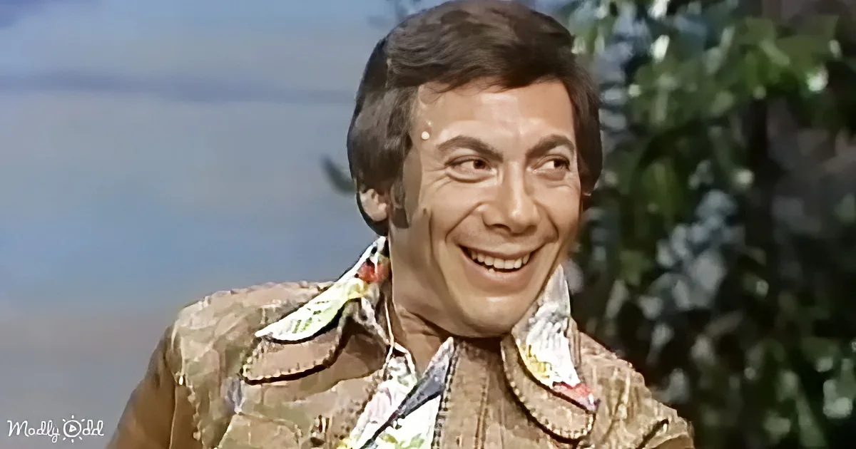 A man in a patterned shirt and jacket smiles, with a blurred background of greenery and sky.
