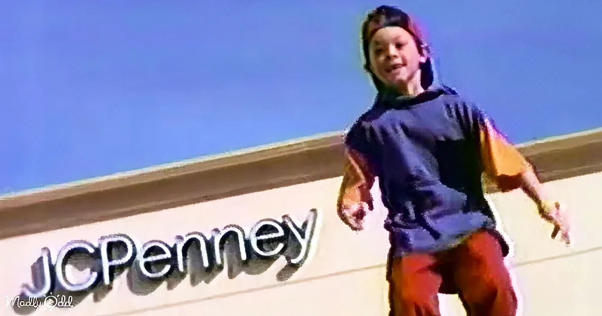 A child in a blue shirt and red pants stands in front of a JCPenney sign under a clear blue sky.