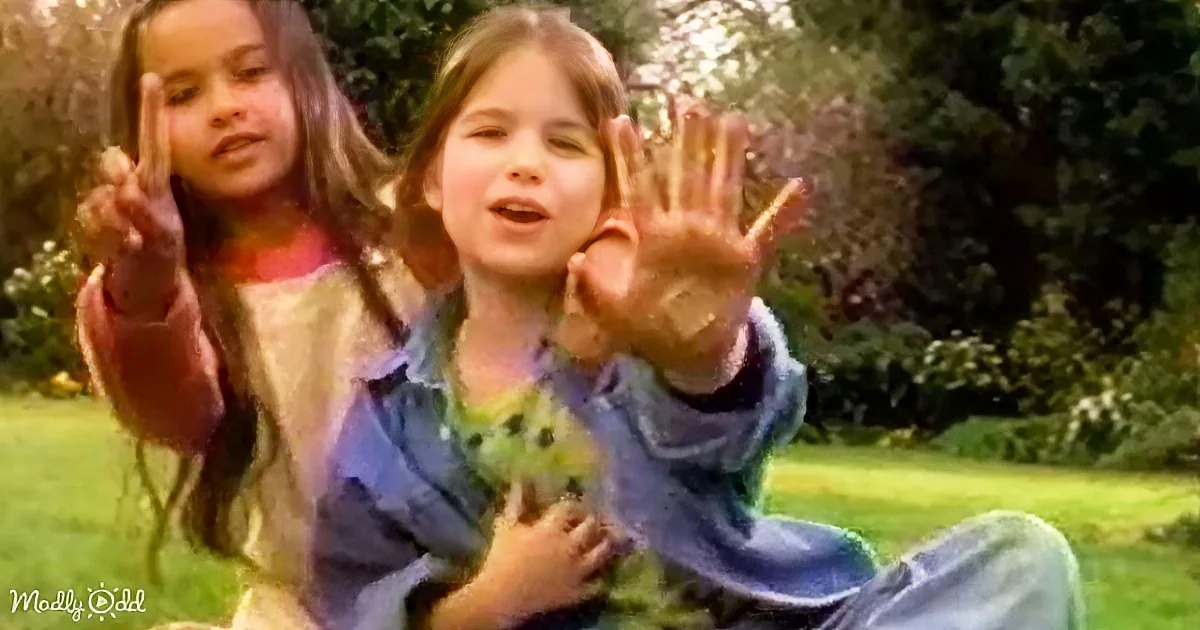 Two children in a park, one making a peace sign, the other waving, both smiling.