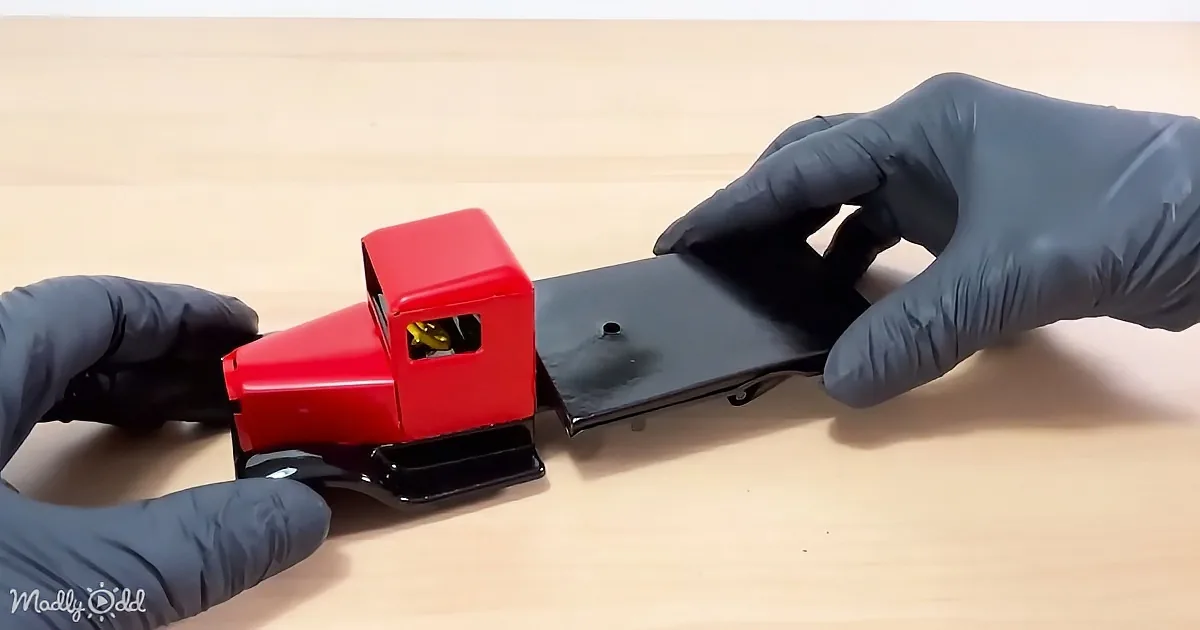 A person in black gloves holds a red toy truck with a black trailer on a wooden surface.