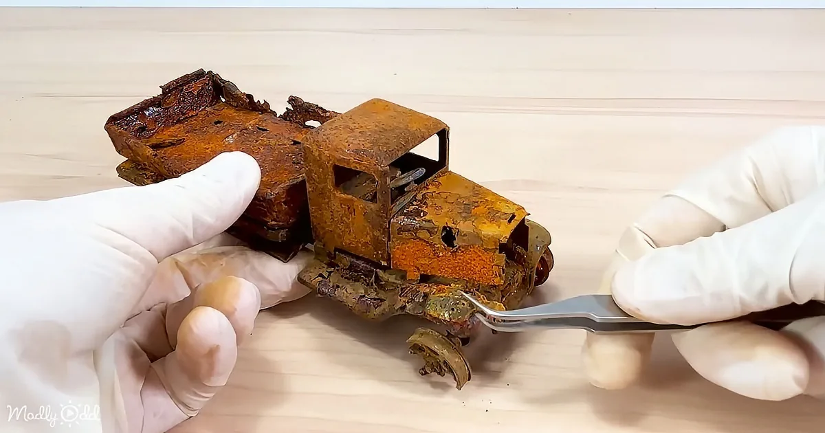 A rusted toy truck is being examined by gloved hands with a tool nearby.