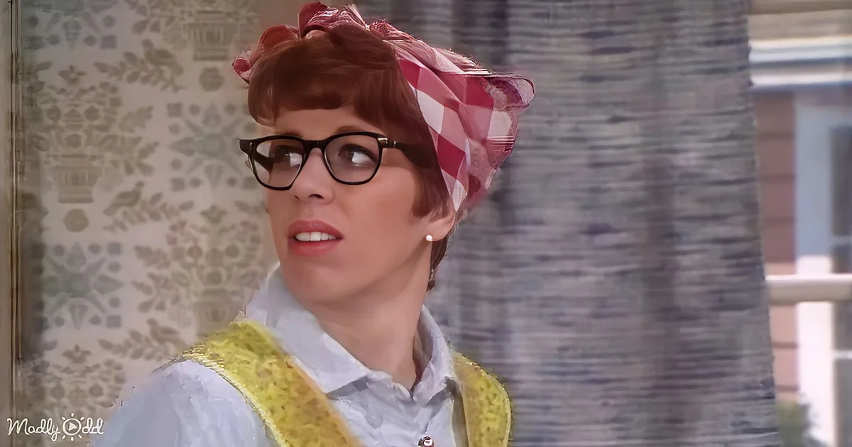 A person with short red hair, wearing glasses, a red checkered headscarf, and a yellow vest over a white shirt.