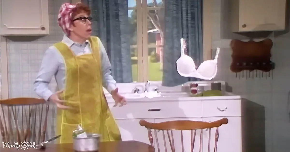 A woman in a yellow apron stands in a kitchen, looking surprised as a white bra floats in the air.