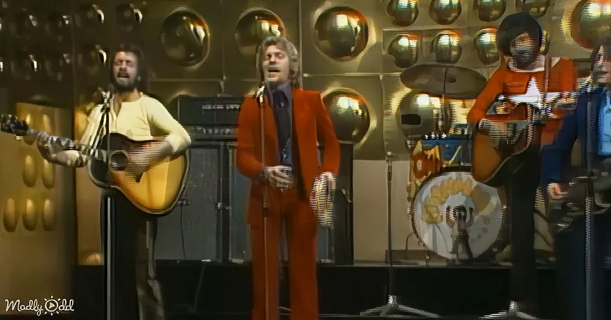 A man in an orange suit sings into a microphone on stage with musicians and a drum set.