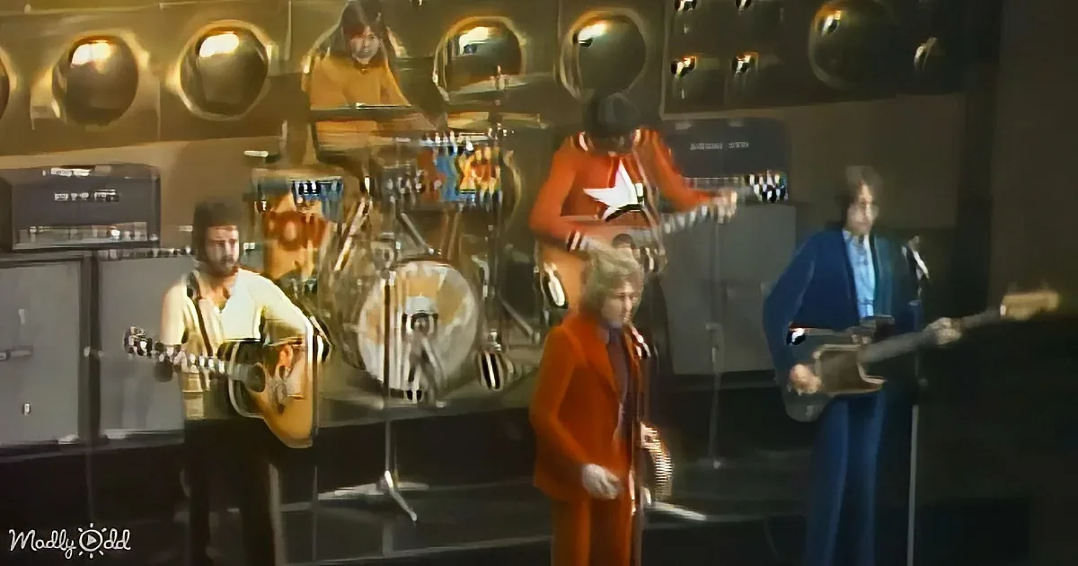 A man in a red suit sings on stage with a band, including a drummer and guitarists.