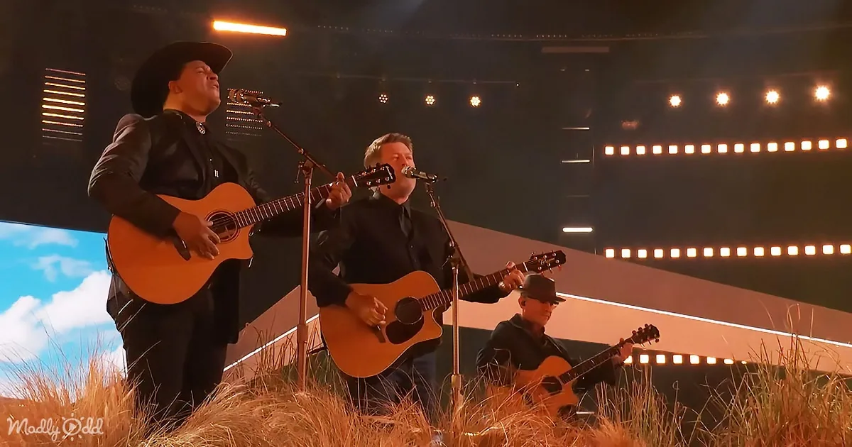 A man in a cowboy hat plays guitar on stage with two other musicians, surrounded by hay and a scenic backdrop.