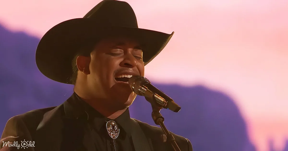 A person in a black cowboy hat and shirt sings into a microphone against a colorful, blurred background.