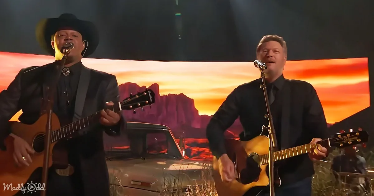 Two men in suits play guitars and sing in front of a sunset backdrop.