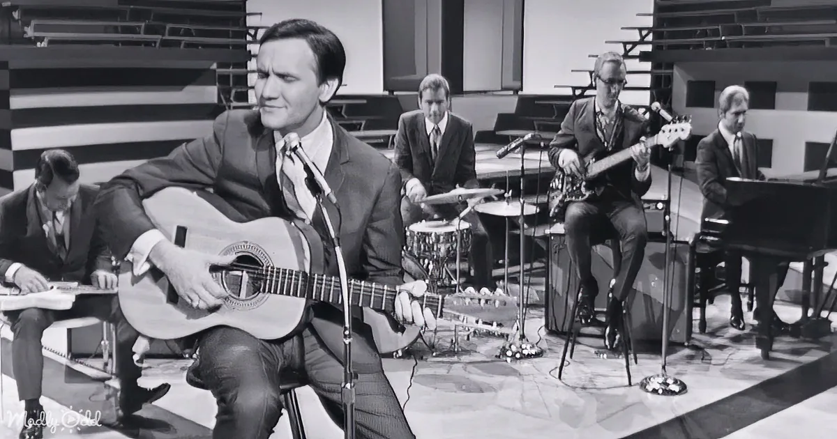A man in a suit plays guitar on stage with a band, including a drummer, bassist, and pianist.