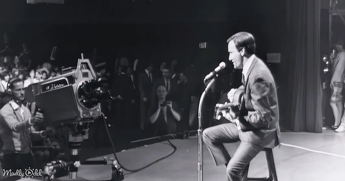 A man in a suit sings into a microphone on stage, with a camera operator and audience in the background.