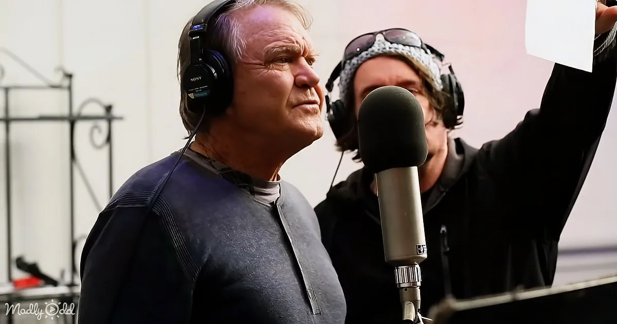 An older man wearing headphones stands in front of a microphone, seemingly recording or speaking.