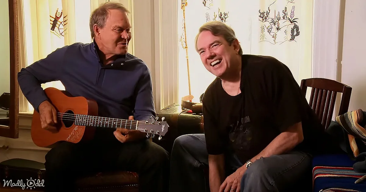 Two men are sitting, one playing a guitar, both smiling, in a cozy room with a window and curtains.