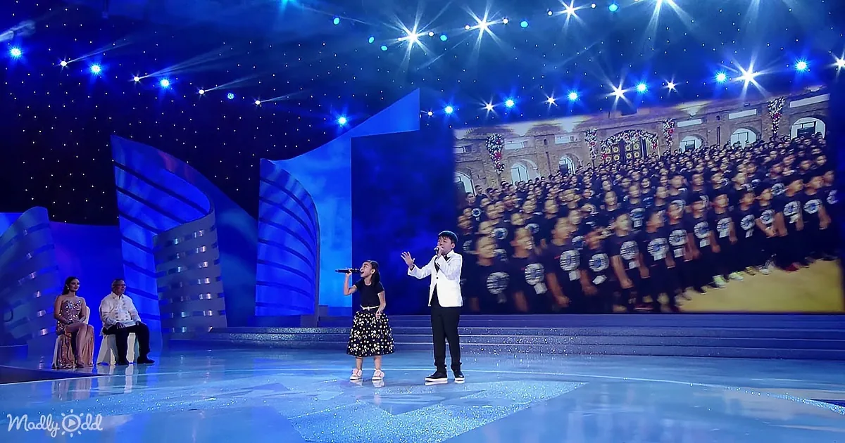 A young girl and boy sing on stage, with a large audience displayed on a screen behind them.