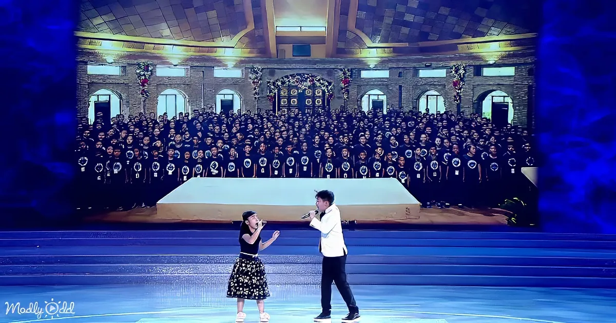A young girl and man sing on stage, with a large choir in matching shirts behind them.
