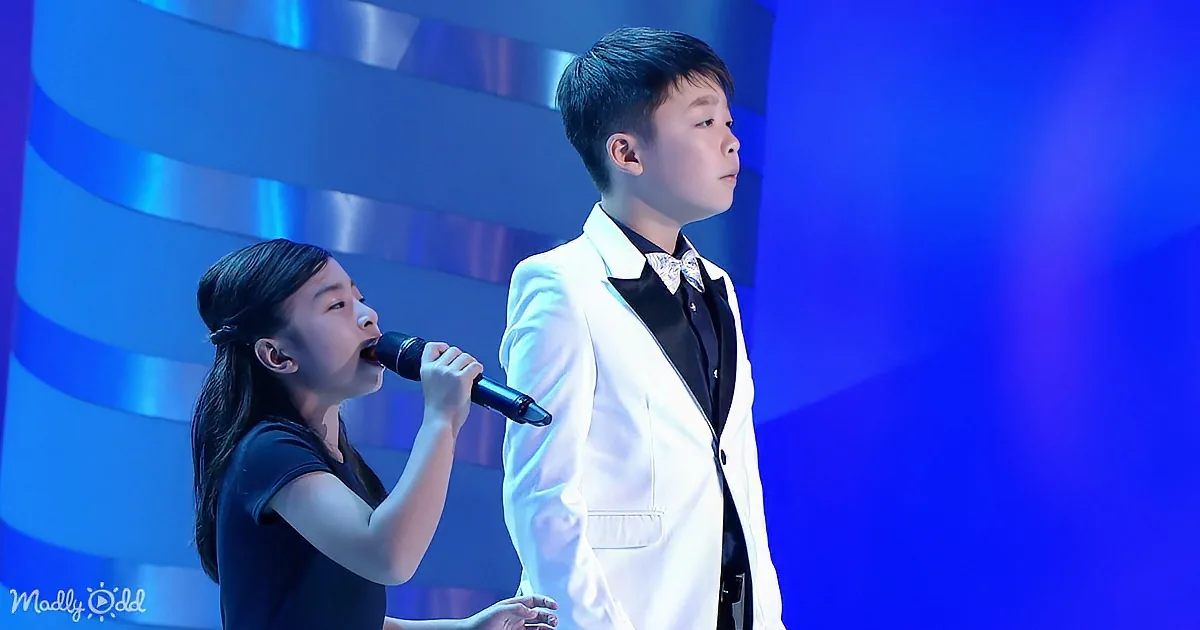 A young girl sings into a microphone while a boy in a white suit stands beside her on stage.