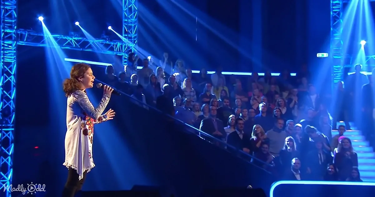 A woman sings on stage under blue lights, surrounded by an audience.