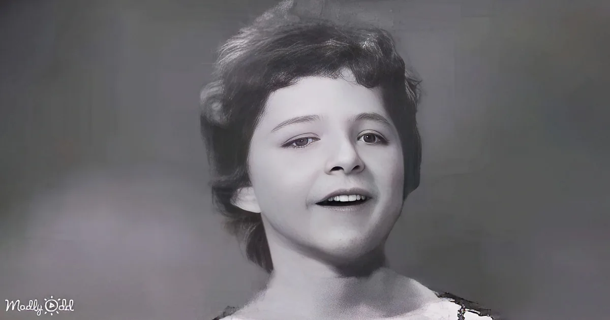 A young child with short hair, smiling, and wearing a white top, set against a plain background.