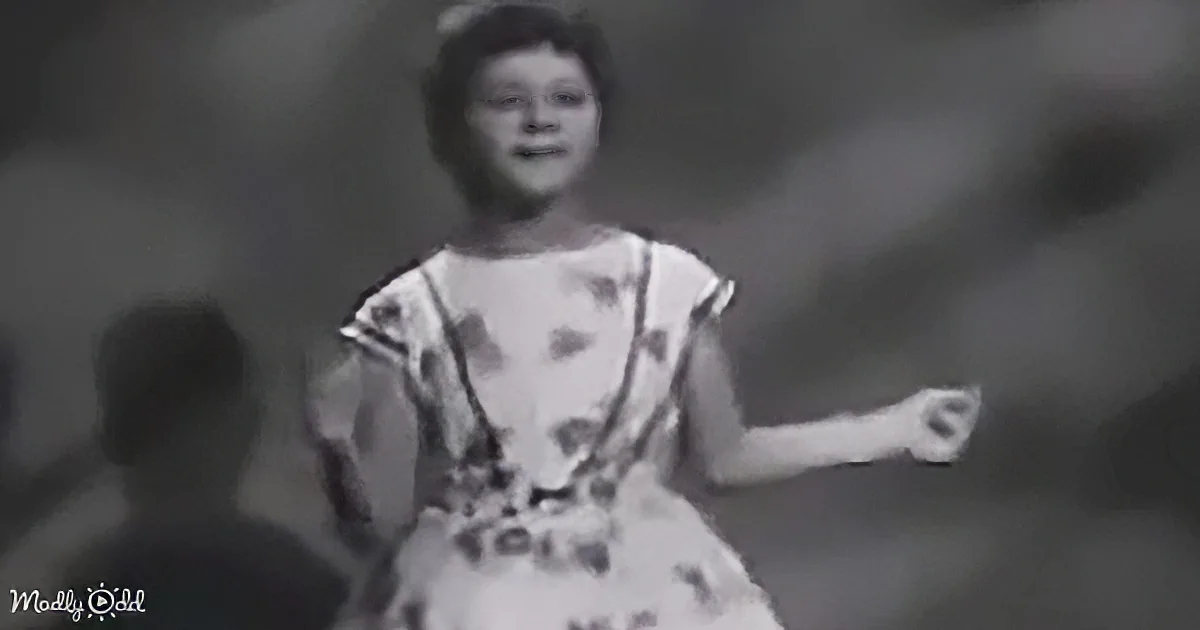 A young girl in a polka-dot dress smiles, arms outstretched, in a black-and-white image.