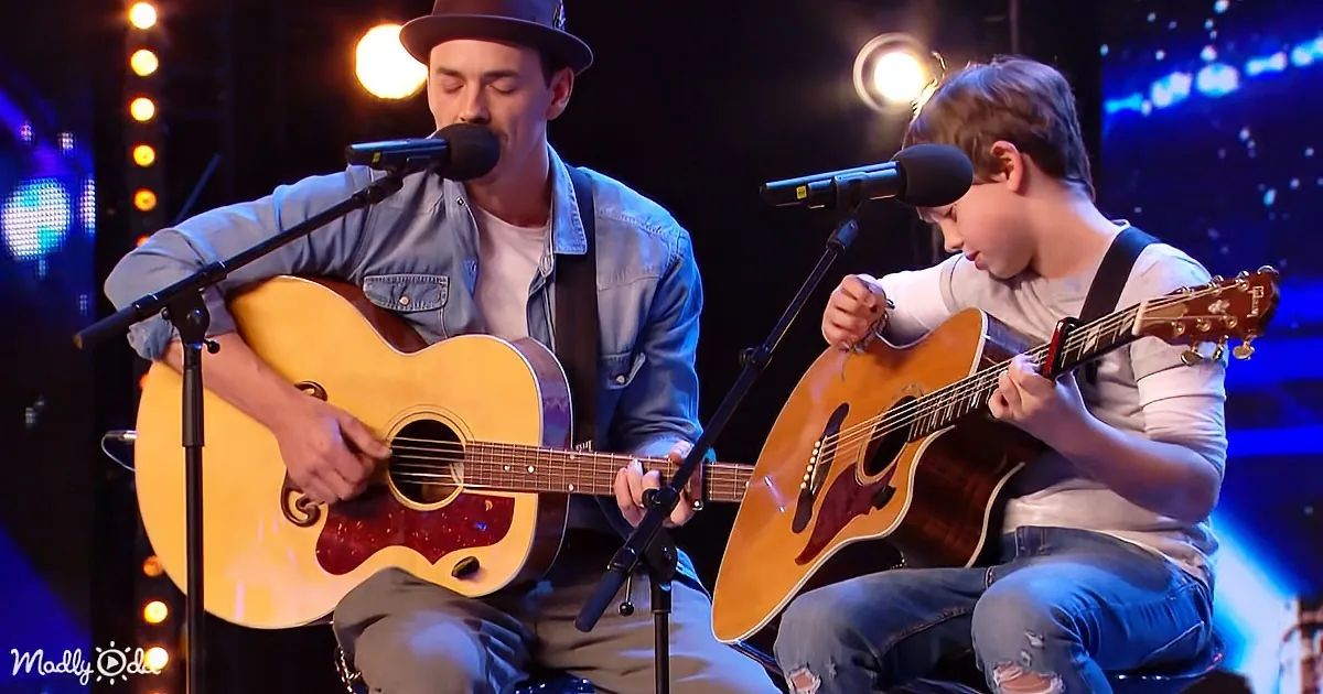 A man and a boy play acoustic guitars on stage, both singing into microphones.