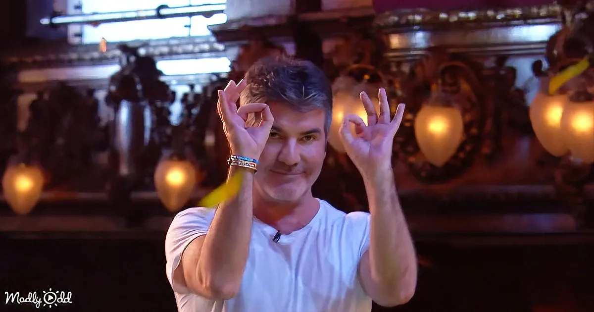 A man in a white shirt makes an "OK" sign with both hands in front of ornate, lit-up background.