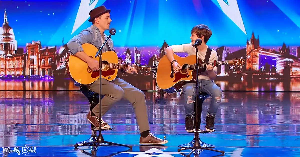 Two musicians play guitars on stage with a cityscape backdrop, one wearing a hat.