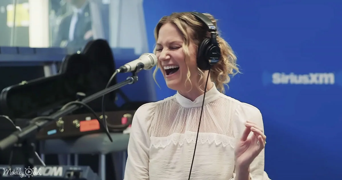 A woman in a white blouse sings into a microphone, wearing headphones, in a recording studio.