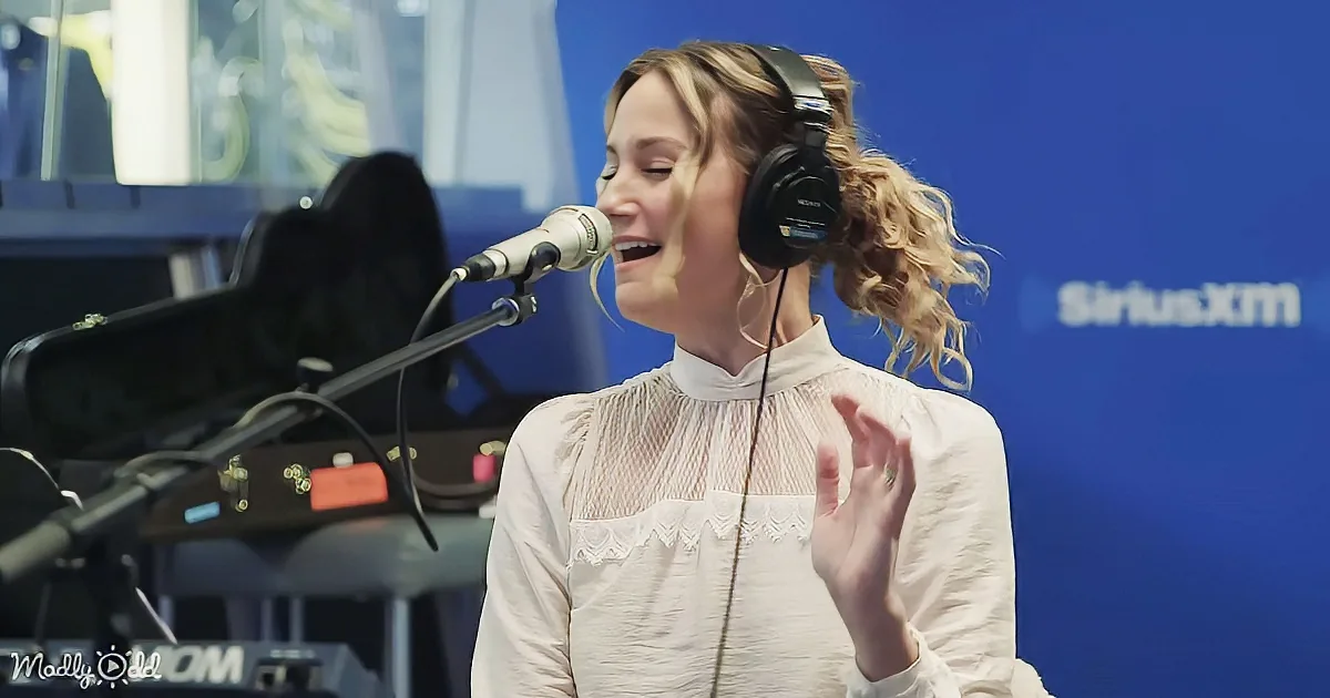 A woman in a white blouse sings into a microphone, wearing headphones, with a guitar case and blue background visible.