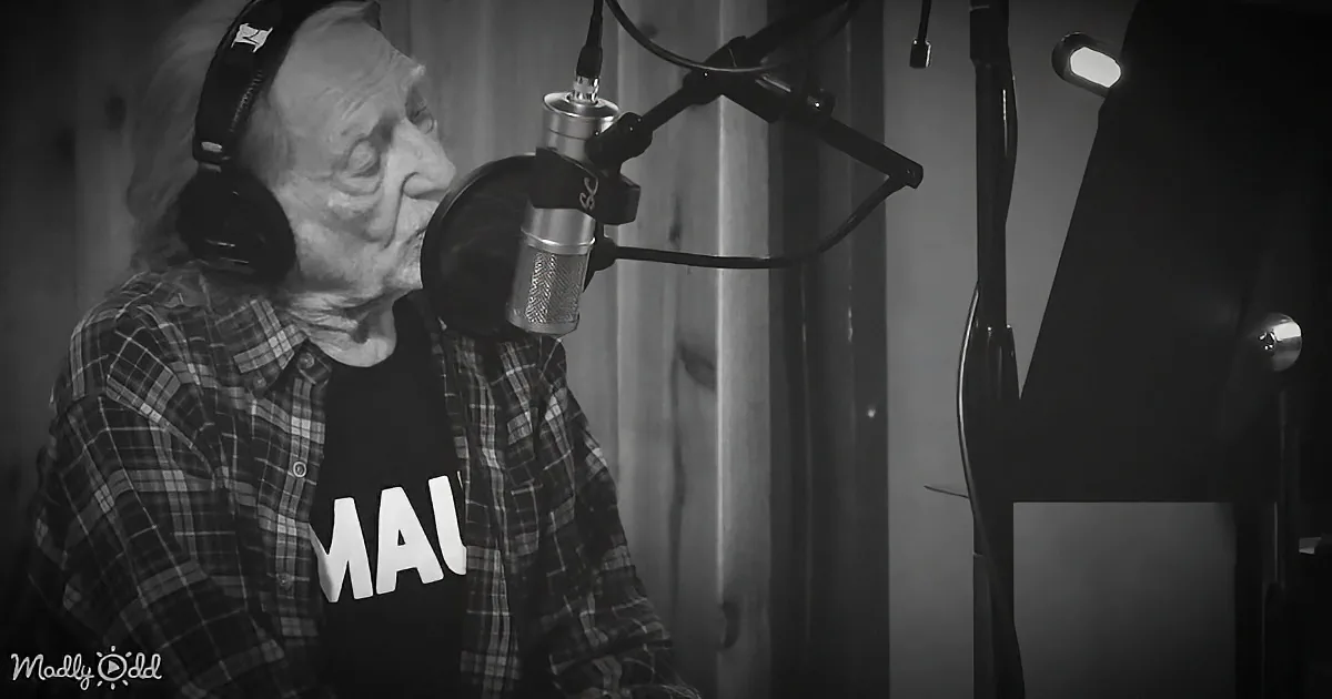An elderly man wearing headphones and a plaid shirt sings into a microphone in a recording studio.