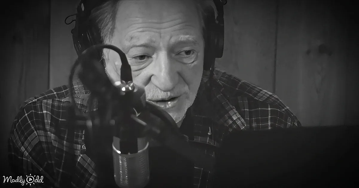 An older man in a plaid shirt records in a studio with headphones and a microphone.