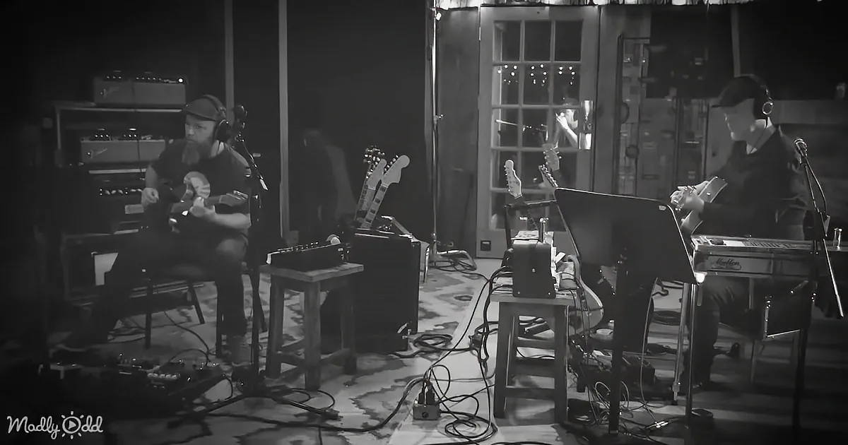 Two musicians play instruments in a dimly lit room with amplifiers and cables.