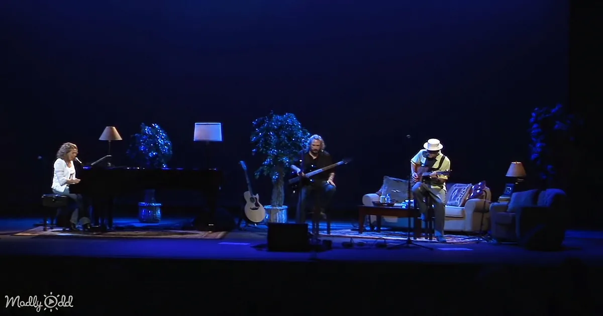 A musician plays piano while another plays guitar on a stage with blue lighting and plants.