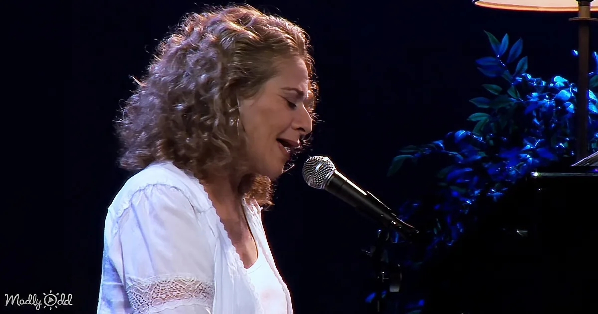 A woman with curly hair sings into a microphone, wearing a white blouse, with a plant and lamp in the background.