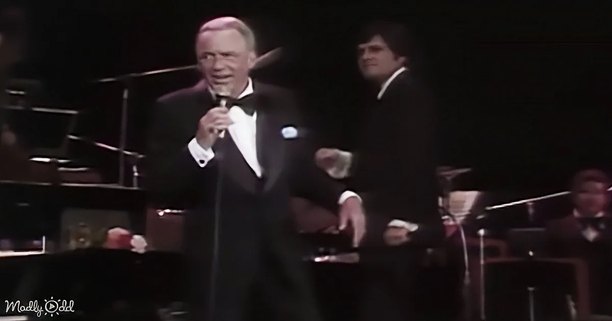 A man in a tuxedo sings into a microphone, accompanied by another man in a suit, with an orchestra in the background.