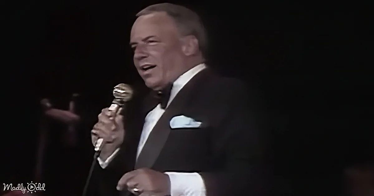 A man in a tuxedo sings into a microphone on stage, illuminated against a dark background.