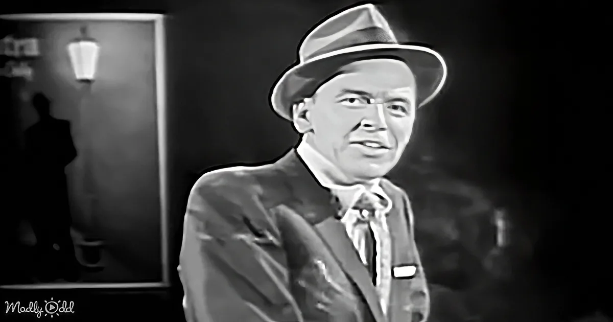 A man in a suit and fedora, wearing glasses, stands in a dimly lit room with a lamp in the background.